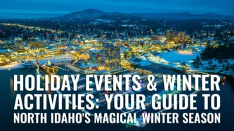 Aerial view of downtown Coeur d'Alene Idaho illuminated at winter dusk with snow-covered cityscape, waterfront resort district, and mountain backdrop