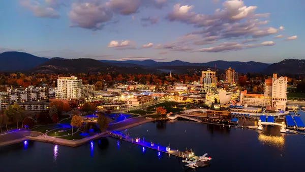 Aerial view of downtown Coeur d'Alene waterfront at sunset with illuminated blue boardwalk, Lake Coeur d'Alene marina, resort district, and mountain backdrop during fall foliage season in North Idaho