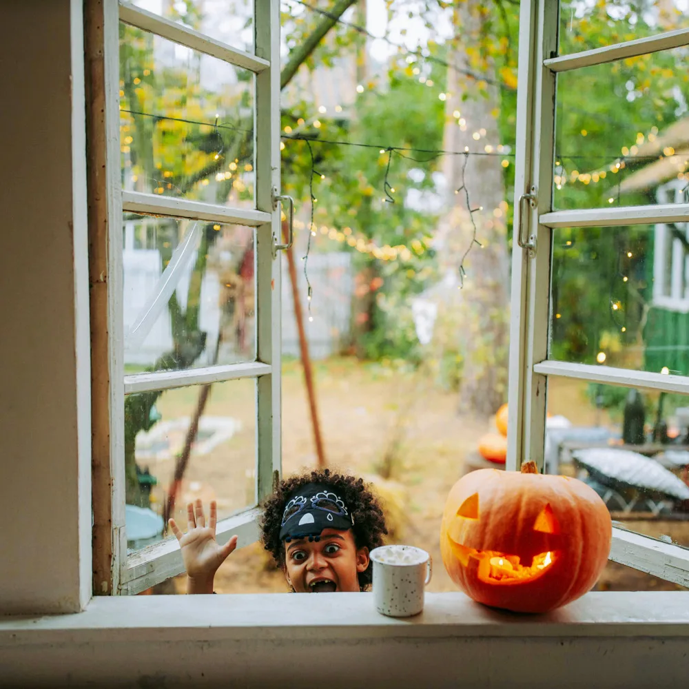 Open window with string lights; a carved jack-o’-lantern on the sill as a child smiles and waves outside.